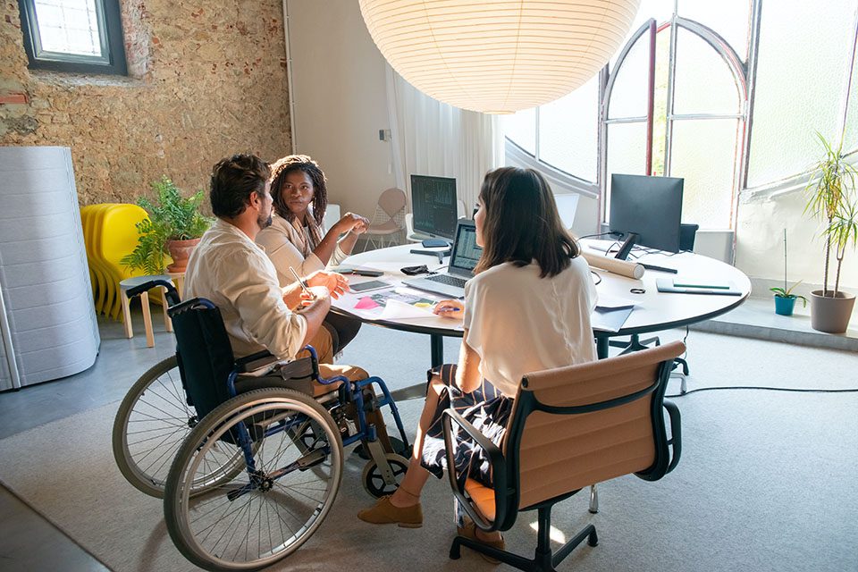 A disabled man in a wheelchair is working with others at a table with computers. Digiboost can assist you with becoming Section 508 compliant.